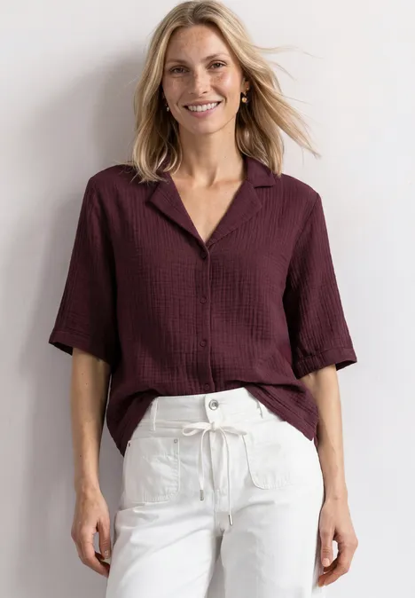 Une femme souriante porte un chemisier bordeaux ample et un pantalon blanc décontracté.