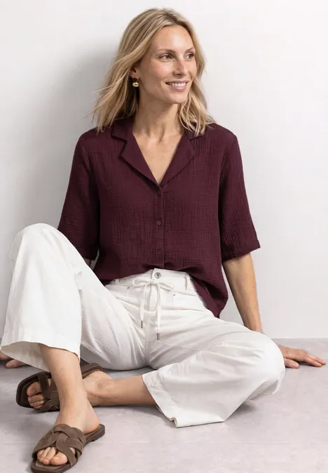 Femme souriante en pantalon blanc ample et chemisier bordeaux à manches trois-quarts, assise détendue sur le sol.