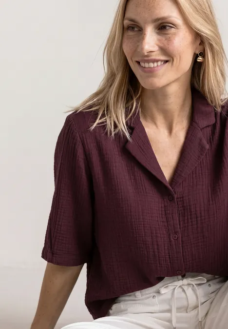Femme aux cheveux blonds, souriante, vêtue d'une blouse bordeaux à poches et manches mi-longues.