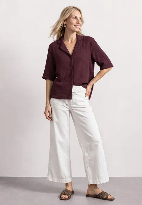 Femme en blouse bordeaux, ample pantalon blanc et sandales marron, souriante devant un fond uni.