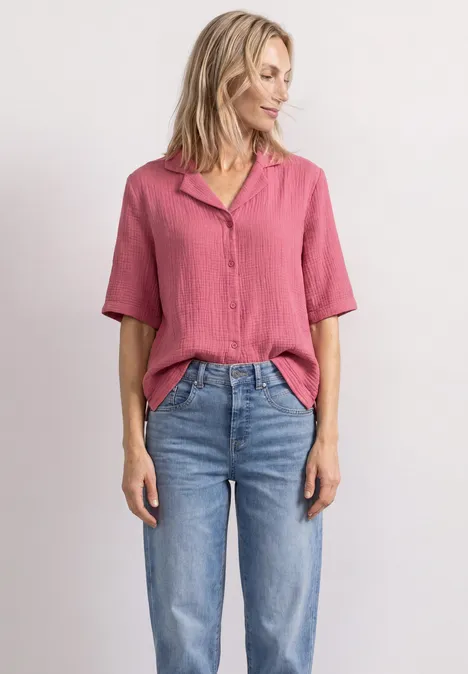 Mousselineblouse met bowlingkraag blush berry Frau in rosa Bluse und blauen Jeans steht lässig vor weißem Hintergrund.