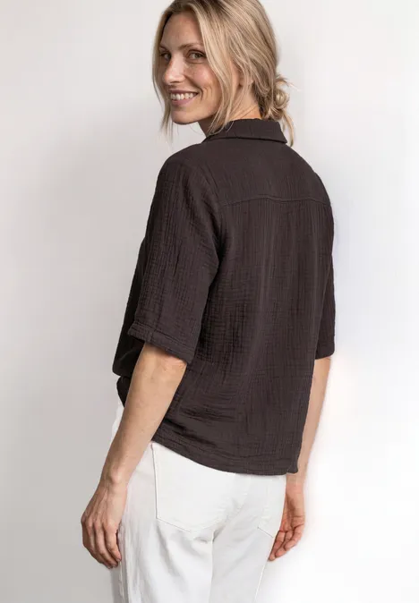 Mousselineblouse met bowlingkraag black coffee Frau mit hellem Haar trägt locker sitzendes, braunes Hemd und weiße Hose, schaut über die Schulter und lächelt.