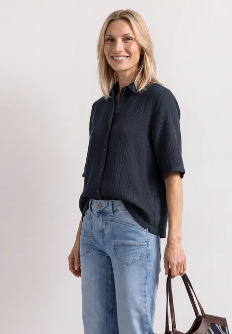Eine lächelnde Frau in dunkler Bluse und Jeans hält eine Tasche.