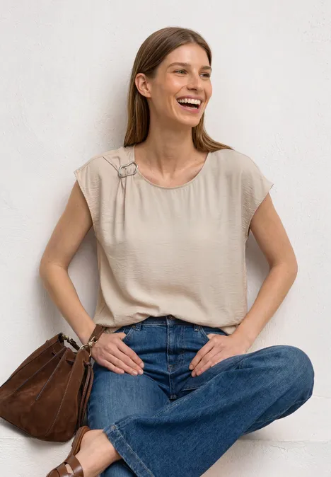 Frau sitzt vor heller Wand, trägt beiges Top, blaue Jeans und braune Tasche.