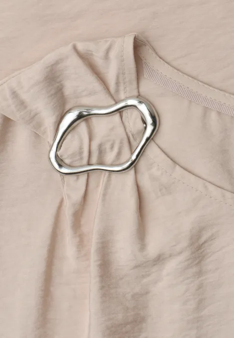 Nahaufnahme eines beigen Oberteils aus zartem Stoff mit unregelmäßigem silbernem Ring an der Schulterpartie.