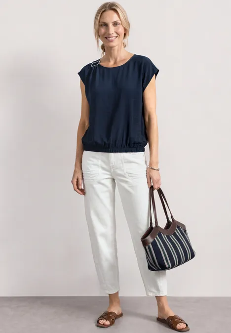 Frau in marineblauer Bluse, weißen Hosen, flachen Sandalen und gestreifter Tasche vor hellem Hintergrund.