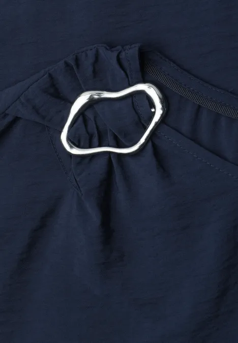 Dunkelblauer Stoff mit silbernem, organisch geformtem Ring als Zierdetail an einer Raffung.