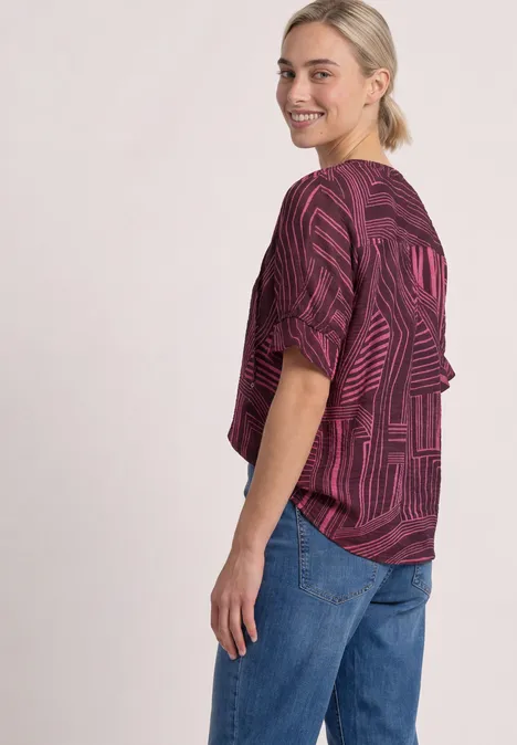 Frau in seitlicher Pose trägt locker geschnittenes lila Muster-Shirt und Jeans, sie lächelt.