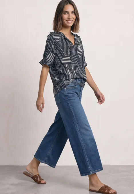 Frau in gemusterter Bluse, weiten Cropped-Jeans und flachen Sandalen vor neutralem Hintergrund.