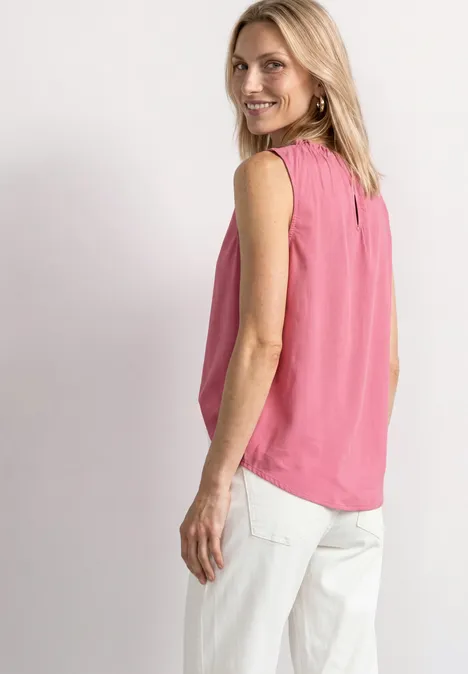 Blonde Frau in ärmelloser rosa Bluse und weißer Hose, seitlich stehend, lächelnd zur Kamera blickend.