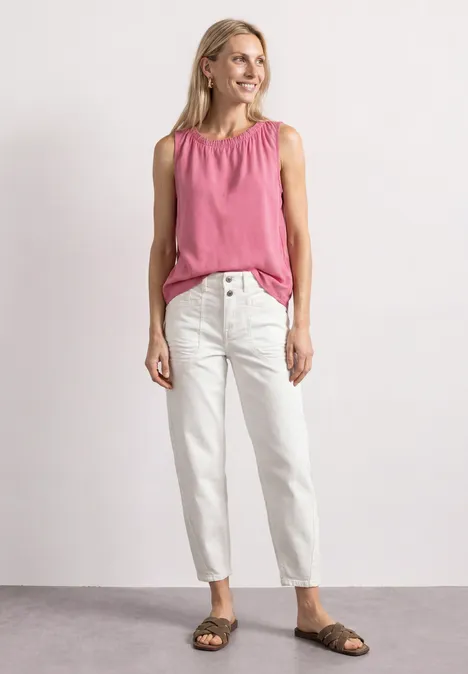 Frau in pinkem ärmellosem Top, weißen Jeans und braunen Sandalen steht lächelnd vor hellem Hintergrund.
