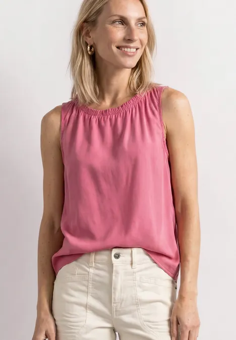 Frau trägt rosa ärmellose Bluse und weiße Jeans, lächelt freundlich.