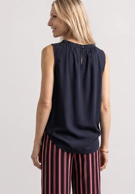 Blouse met gerimpelde ronde hals darkest blue Frau von hinten, trägt dunkelblaues ärmelloses Top mit gerafftem Kragen und rot-blaue, vertikal gestreifte Hose.