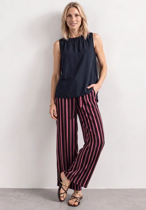 Blouse met gerimpelde ronde hals darkest blue Frau in schwarzem Top, rot-schwarz gestreiften Hosen und Sandalen posiert vor heller Wand.