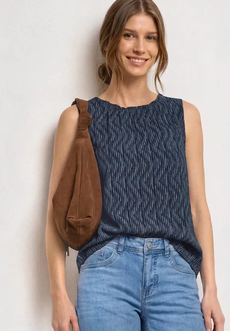Femme en top en maille bleu foncé texturé et jean, portant un sac marron épaule, devant un mur blanc.