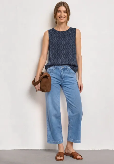 Une personne porte un top sans manches bleu, un jean ample, des sandales marron et tient un sac assorti.
