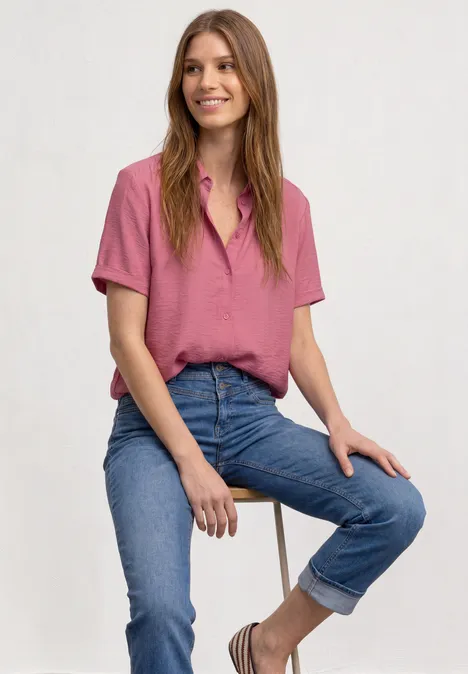 Eine lächelnde Frau sitzt auf einem Hocker, trägt rosa Hemd und hochgekrempelte blaue Jeans.