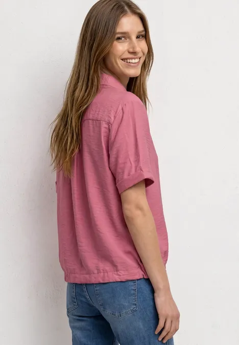 Eine Frau in rosa Kurzarmbluse und Jeans blickt lächelnd über die Schulter vor weißem Hintergrund.