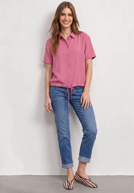 Lächelnde Frau in rosa Hemdbluse, locker sitzender Jeans mit aufgekrempelten Beinen und Sandalen vor weißer Wand.