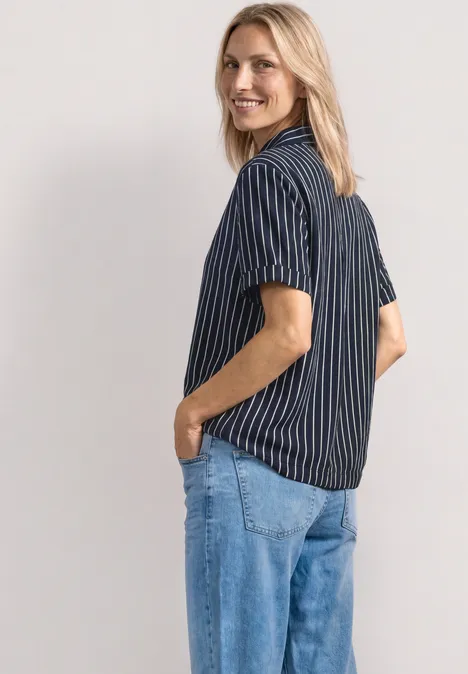 Personne vue de dos en trois-quarts, chemise rayée bleu et blanc, jeans clairs, mains dans les poches.