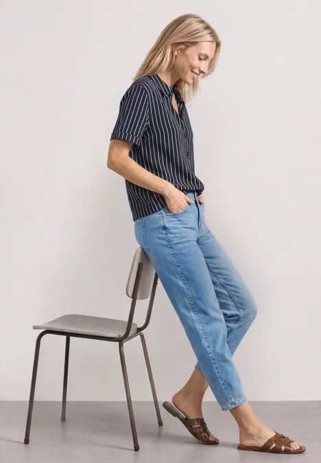 Une femme décontractée en chemise rayée, jean bleu et sandales s'appuie sur une chaise.