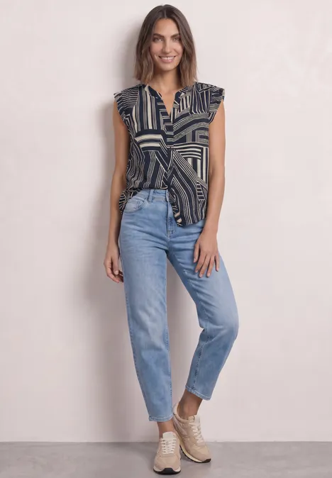Een glimlachende vrouw draagt een blouse met patroon, opgerolde jeans en beige enkellaarzen voor een lichte achtergrond.