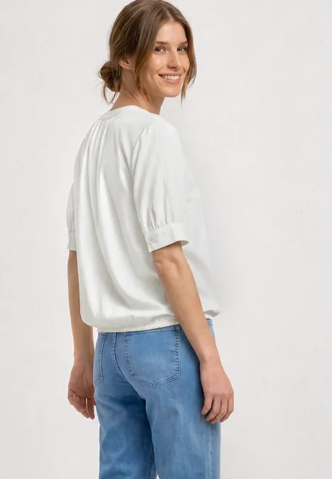 Blouse met korte mouwen, split in de hals en elastische zoom off white Vrouw in los wit t-shirt en blauwe jeans, van achteren schuin gefotografeerd.