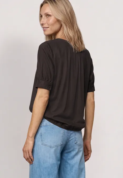 Frau steht mit Blick nach hinten, trägt schwarzes T-Shirt mit hochgekrempelten Ärmeln und helle Jeans.