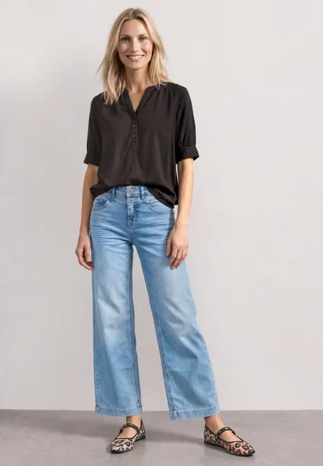 Eine blonde Frau in schwarzem Hemd, hellblauer Jeans mit weitem Bein und gemusterten Sandalen steht vor heller Wand.