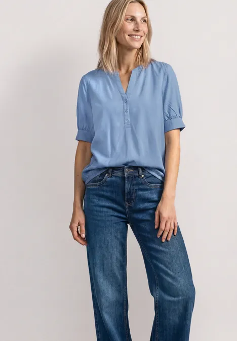 Blouse met korte mouwen, split in de hals en elastische zoom northern blue Vrouw in lichtblauw bloesje en blauwe spijkerbroek voor lichte achtergrond.