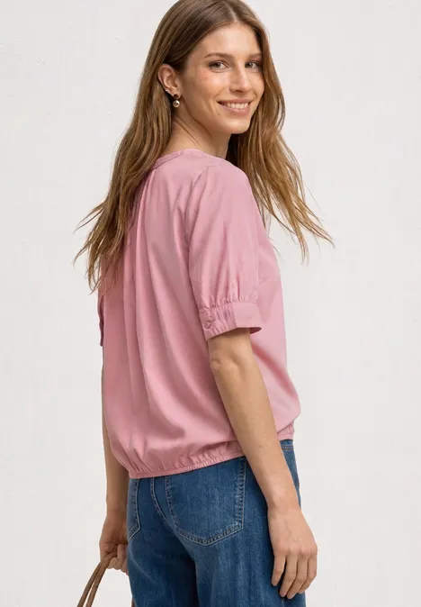 Blouse met korte mouwen, split in de hals en elastische zoom vintage blush Een vrouw van achteren met een roze blouse en blauwe jeans, die een tas vasthoudt.