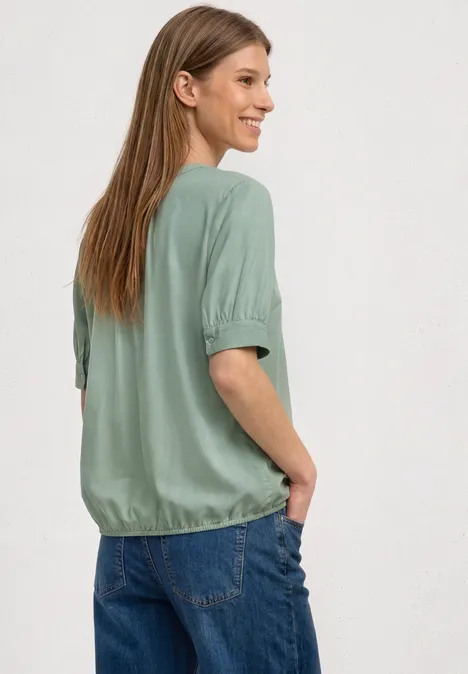 Kurzarm Bluse mit Split Neck und Elastiksaum eucalyptus green Frau mit langen Haaren von hinten, trägt grünes Oberteil und blaue Jeans, Hände in den Taschen.