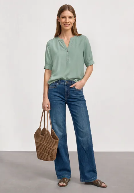 Kurzarm Bluse mit Split Neck und Elastiksaum eucalyptus green Eine Frau in grüner Bluse, blauen Jeans, Sandalen, mit geflochtener Tasche vor neutralem Hintergrund.