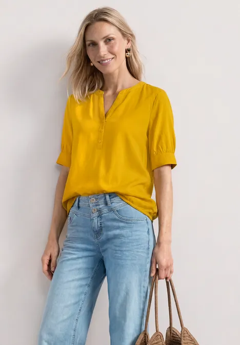 Blouse met korte mouwen, split in de hals en elastische zoom saffron yellow Vrouw in gele blouse en blauwe spijkerbroek met bruine handtas voor een witte achtergrond.
