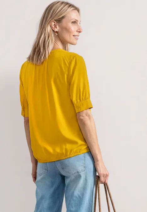 Blouse met korte mouwen, split in de hals en elastische zoom saffron yellow Een vrouw in een geel bloesje en jeans gezien van opzij, ze draagt een handtas.