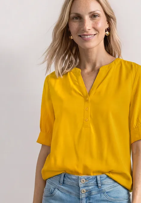 Blouse met korte mouwen, split in de hals en elastische zoom saffron yellow Een vrouw draagt een mosterdgele blouse met V-hals en halflange mouwen, nonchalant ingestopt in een lichtblauwe jeans.