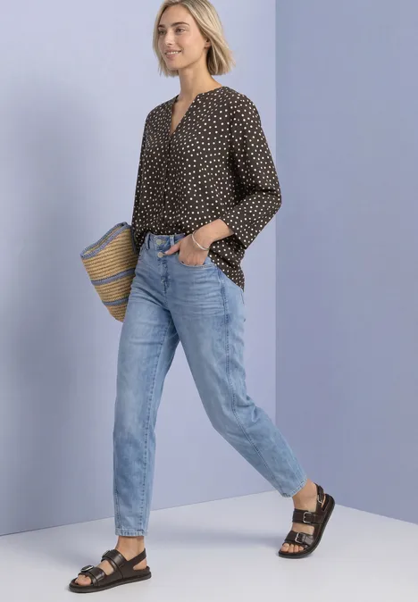 Eine Frau in Jeans, schwarzer gepunkteter Bluse und Sandalen trägt eine Strohtasche vor lila Wand.