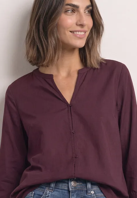 Blouse met 3/4-mouwen en split in de hals rustic rouge Vrouw met bruin haar draagt een bordeauxrode blouse met V-hals en jeans, ze glimlacht terwijl ze opzij kijkt.