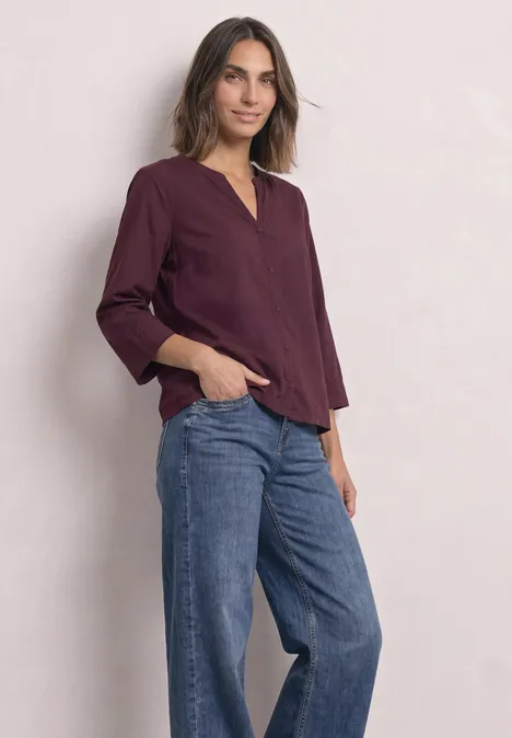 Blouse met 3/4-mouwen en split in de hals rustic rouge Vrouw in bordeauxroze blouse en wijde jeans, ontspannen pose voor een lichte achtergrond.