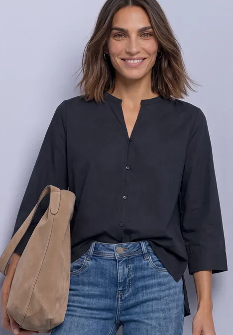 Femme souriante en chemisier noir, jean bleu et sac beige sur fond gris clair.