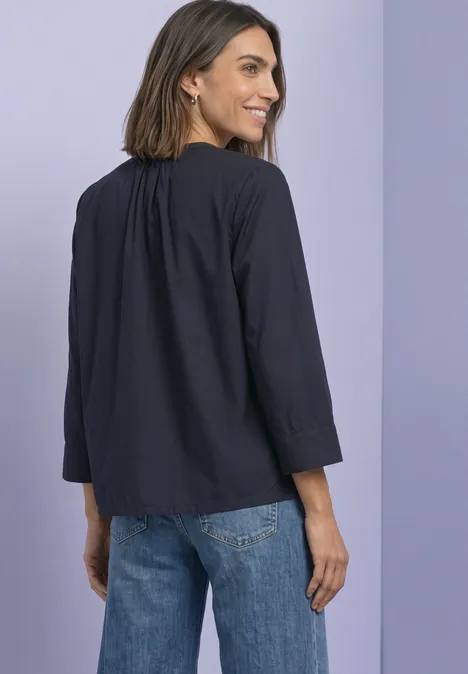 Femme de dos en chemisier bleu foncé et jean, regard par-dessus l'épaule, fond violet clair.