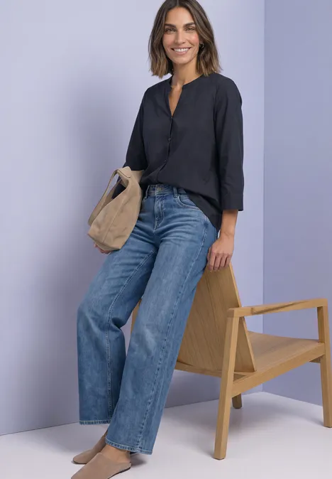 Femme en chemise noire et jean large, sac beige à côté d’un fauteuil sur fond violet.