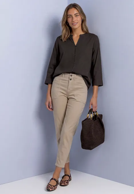 Eine Frau in schwarzer Bluse, beigen Hosen und Sandalen hält eine braune Tasche lächelnd.