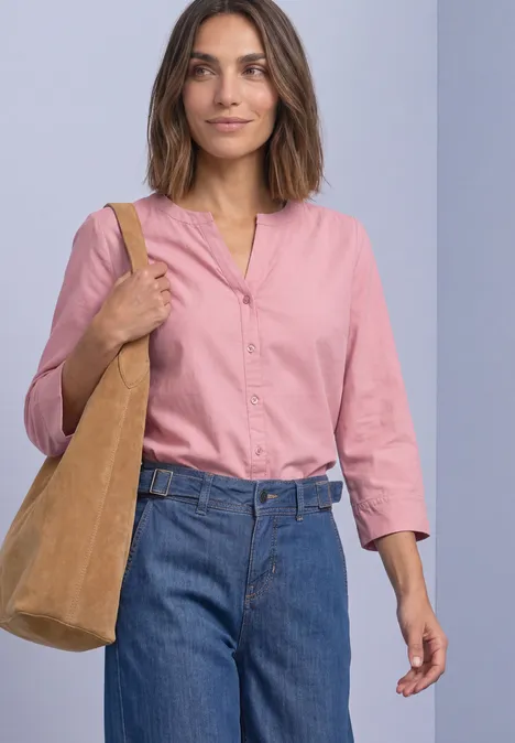 Frau in rosa Bluse, blauen Jeans und beiger Schultertasche vor neutralem Hintergrund.
