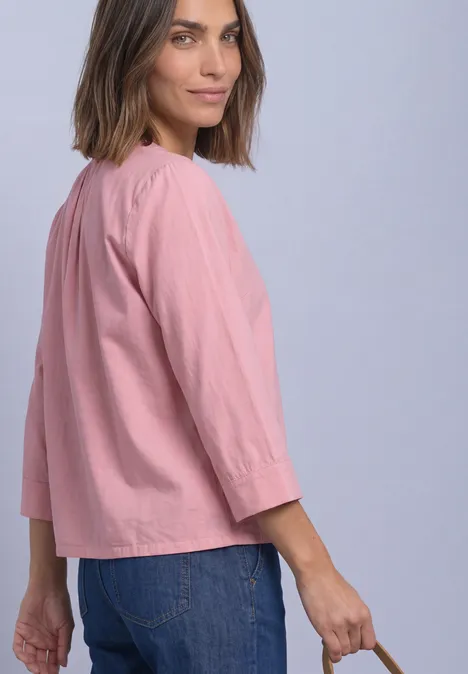 Frau trägt locker sitzende rosa Bluse mit 3/4-Ärmeln und blaue Jeans, Blick über die Schulter.