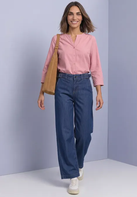 Frau in blauer Jeans, rosa Bluse und weißen Sneakern mit Schultertasche geht vor hellviolettem Hintergrund.