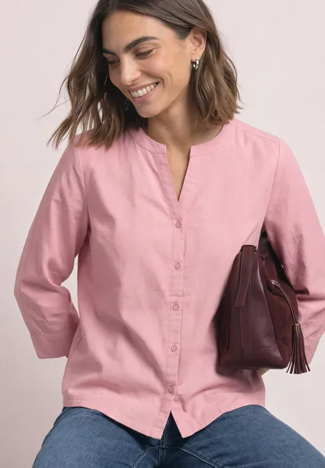 Vrouw lacht, draagt een roze blouse en spijkerbroek en houdt een bruine handtas.
