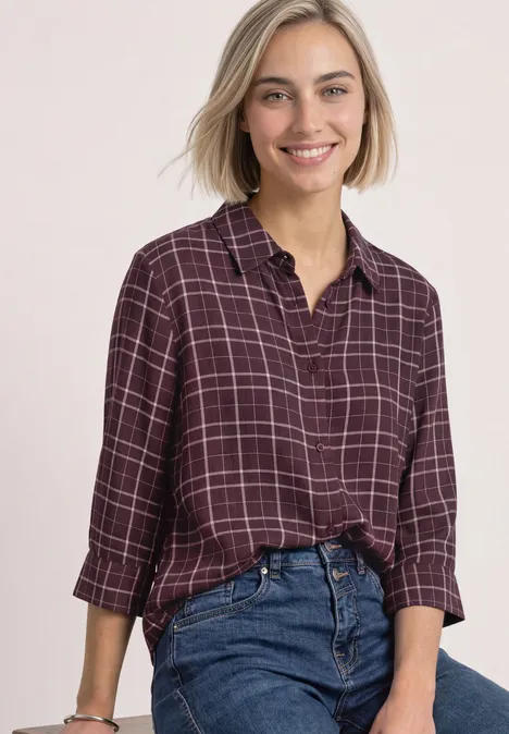 Overhemdblouse met 3/4-mouwen en patroon rustic rouge Eine lächelnde Frau in einem karrierten weinroten Hemd, die locker eine Jeans trägt vor hellem Hintergrund.