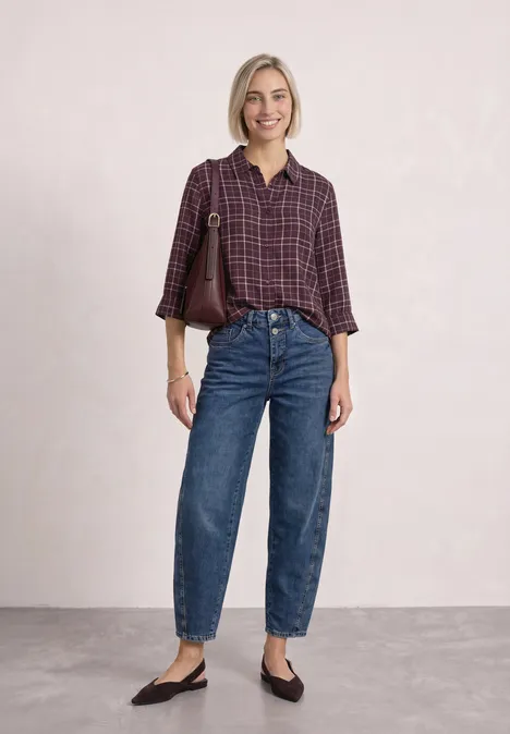 Overhemdblouse met 3/4-mouwen en patroon rustic rouge Eine Frau mit karierter Bluse, hoher Jeans und flachen Schuhen posiert lächelnd mit Schultertasche.