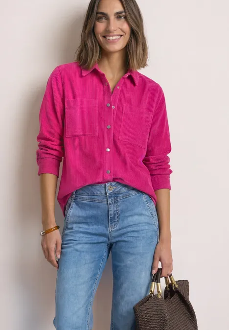 Corduroy overhemd met lange mouwen en knopen magenta dream Frau in pinkem Hemd und blauen Jeans lächelt und hält eine braune Handtasche.